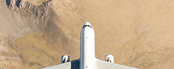 Top-down view of a commercial jet flying over a rugged, sandy landscape, highlighting the aircraft’s wingspan and clean white fuselage.