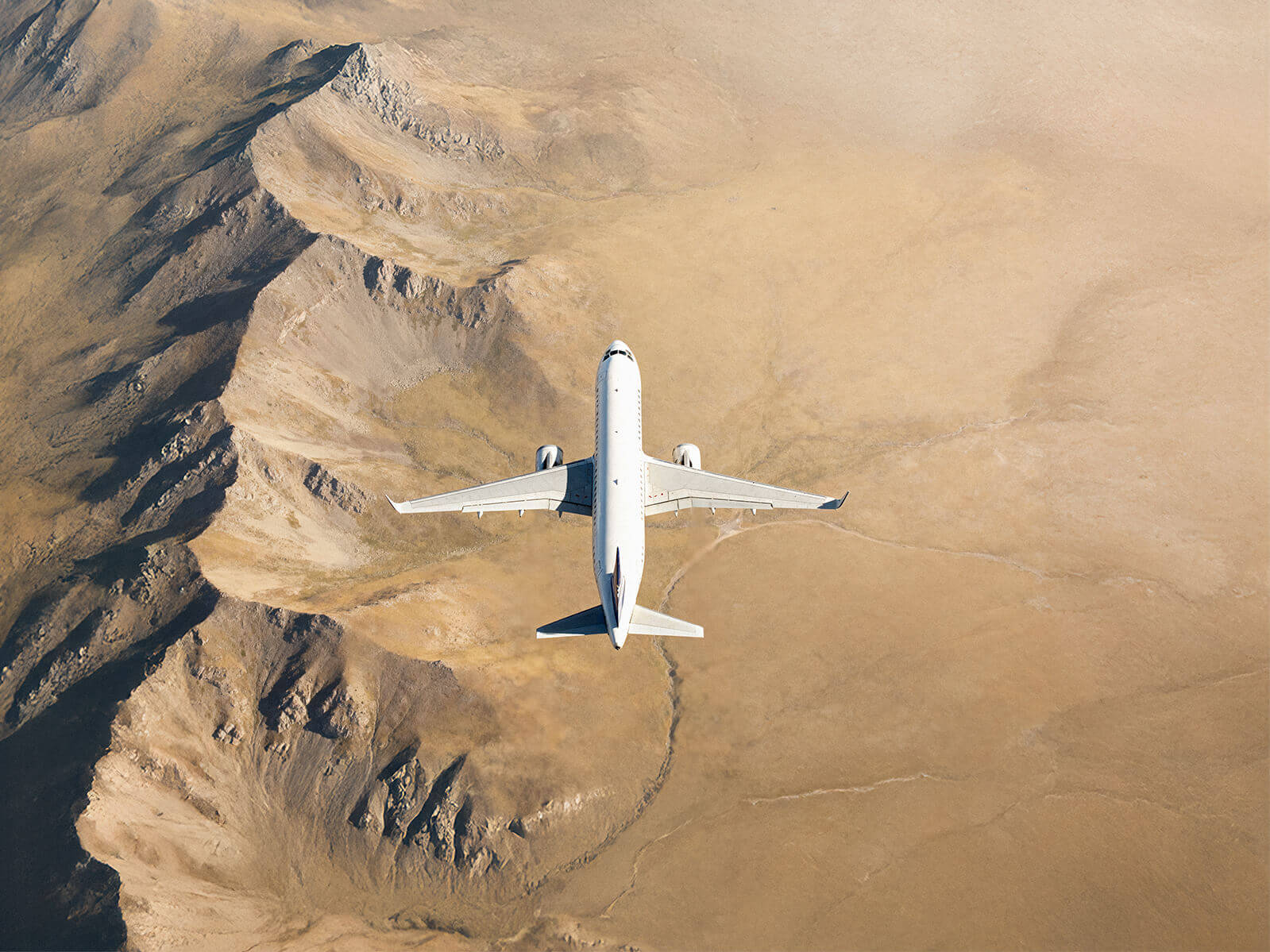 Top-down view of a commercial jet flying over a rugged, sandy landscape, highlighting the aircraft’s wingspan and clean white fuselage.