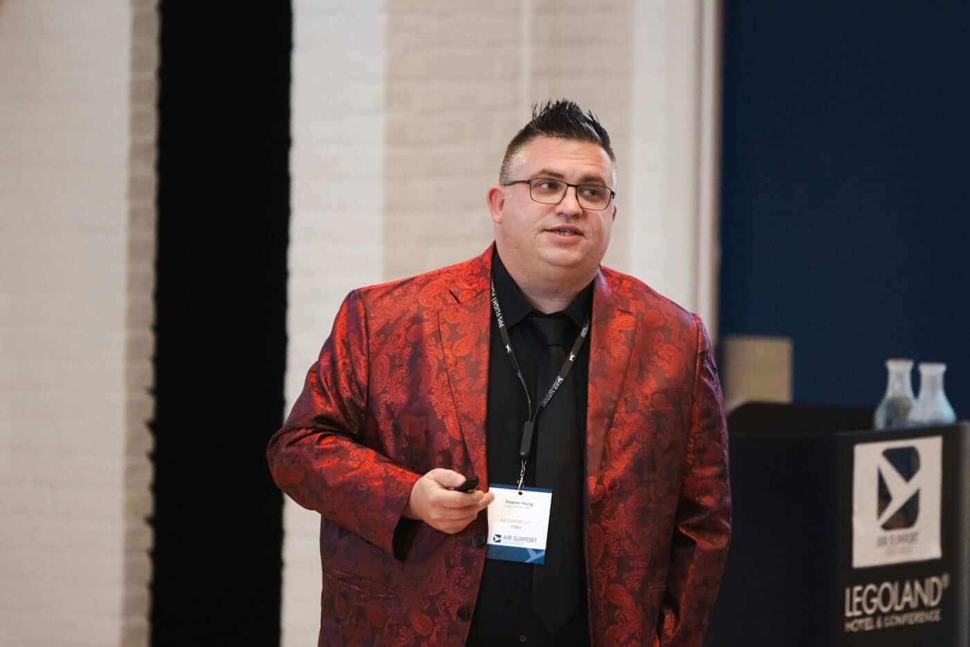 Photo of a speaker presenting at the 2024 User Group Meeting, wearing a striking red patterned jacket and a lanyard with a name tag displaying the AIR SUPPORT logo. The setting includes a podium with the Legoland Hotel & Conference branding.