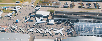 Aerial view of airport apron filled with parked aircraft, visitors walking between planes, exhibition tents, and terminal building nearby.