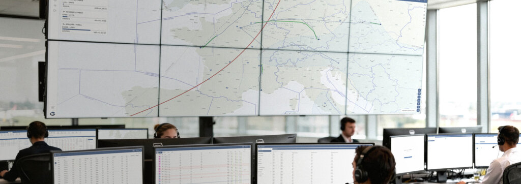 Operations control room with staff at multiple monitors, large wall display showing flight routes and airspace map in real time.